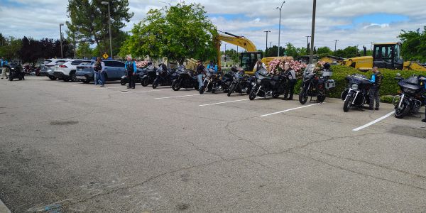 Motorcycles and cars parked with people standing nearby under a partly cloudy sky.