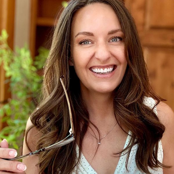 Smiling woman holding glasses and a thick book, sitting indoors.
