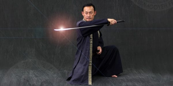 Kummooyeh Gumdo Grand Master Jang kneels in a deep purple silk master uniform while holding a sword.