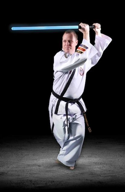 Chief Instructor Patrick Chamberlain wears a white martial arts uniform while holding a combat sabero