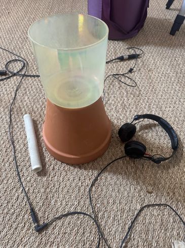 A terracotta pot with a translucent plastic container on top, surrounded by headphones and cables on carpet.