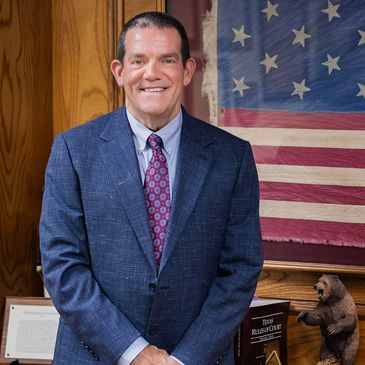 Man in suit standing by an American flag and bear statue.