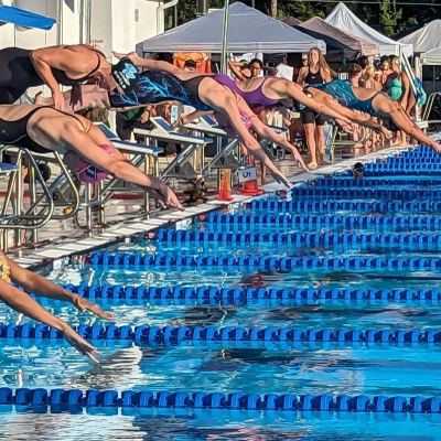 Swimmers diving into the pool at the start of a race in a sunny outdoor competition.
