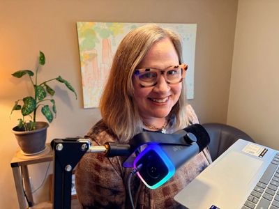 Smiling woman with glasses holding a laptop and a microphone in a cozy room.