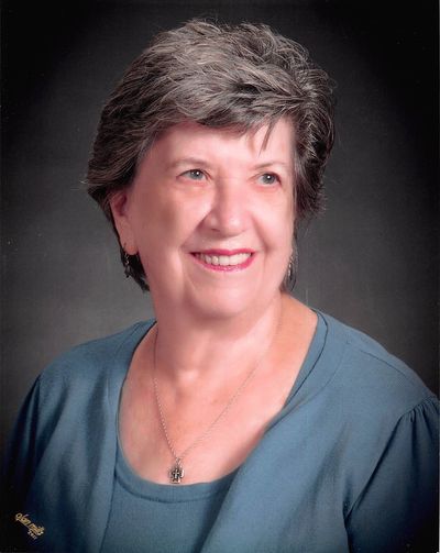 Smiling elderly woman with short gray hair wearing a blue top and cross necklace.