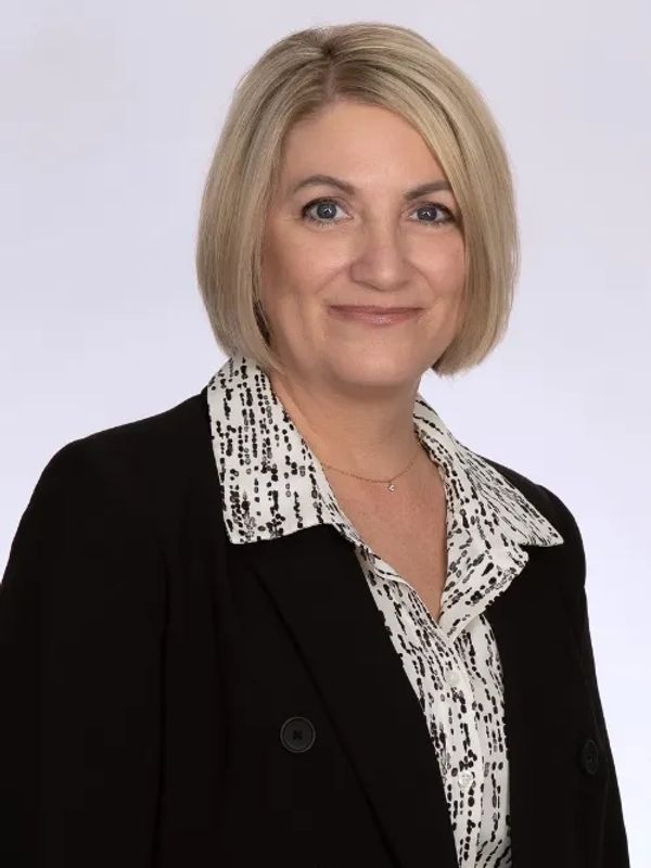 Professional woman with blonde bob haircut wearing a black blazer and patterned blouse.