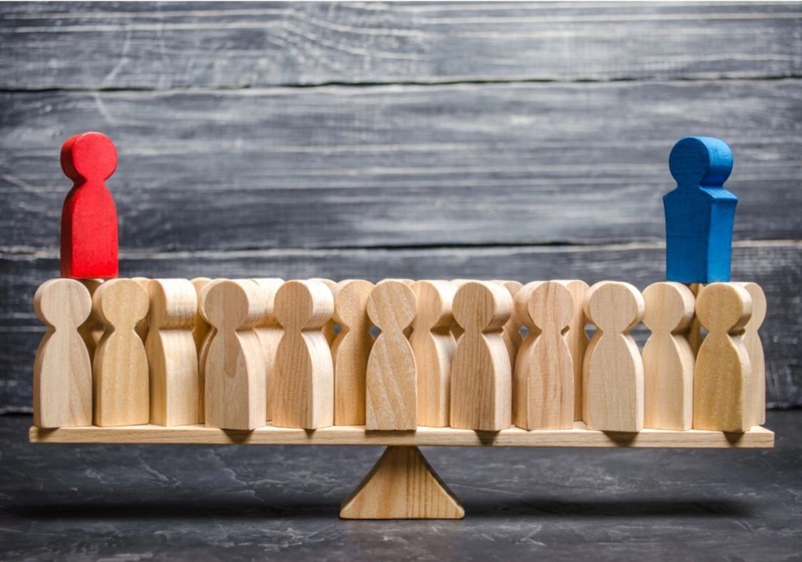 Wooden figures balanced on a seesaw, with red and blue figures standing apart.
