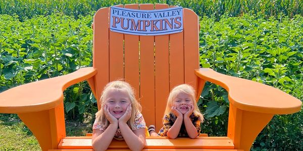 Two children sit on a giant orange chair in a pumpkin patch.