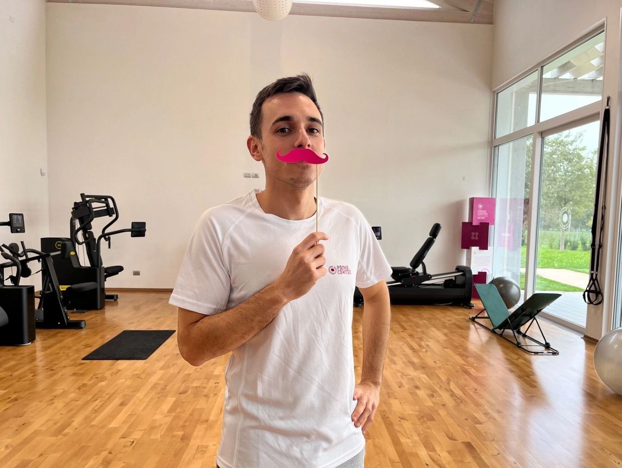 Man in gym holding a pink paper mustache on a stick, posing playfully.
