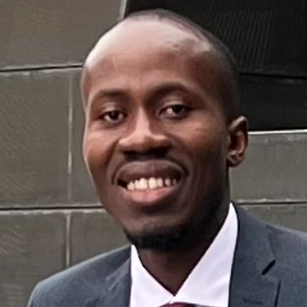 Man in a suit smiling outdoors near a large modern sculpture.