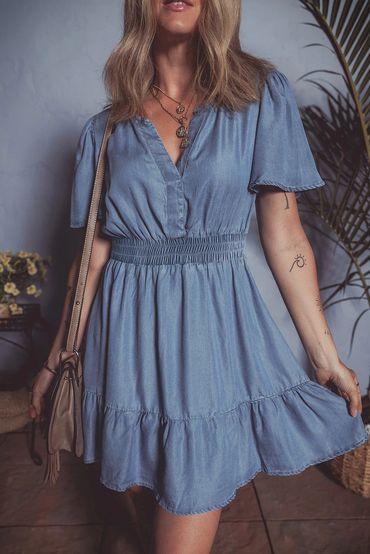 Woman wearing a blue denim dress with layered necklaces and carrying a beige shoulder bag.