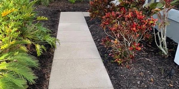 Concrete pathway lined with colorful plants and mulch beside a house.