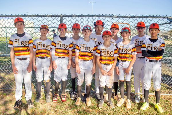 Youth baseball team in matching uniforms posing by chain-link fence.