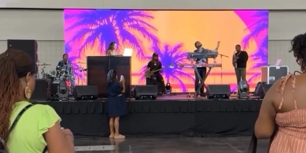 Large LED video wall on stage displaying a palm tree sunset visual behind a live band