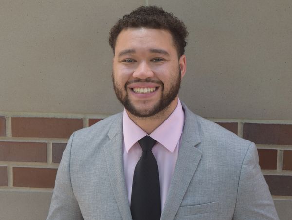 Jordan Colbert, a man with black hair and a grey suit with pink shirt and black tie