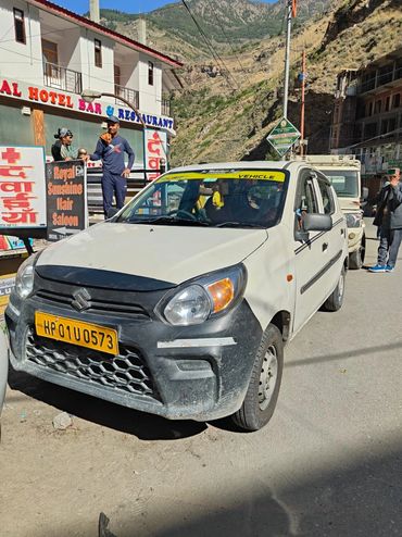 White Suzuki taxi parked on a mountain road near a hotel and hair salon.