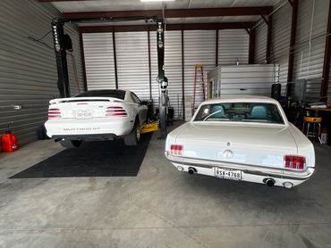 Two white Ford Mustangs parked inside a garage, one on a lift and the other on the floor.