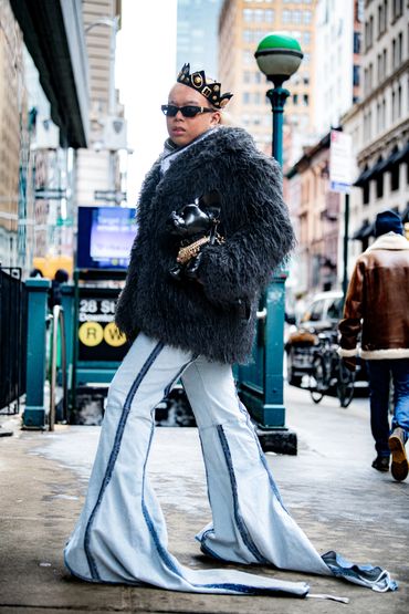 Person wearing oversized flared jeans, a furry black jacket, and a decorative crown on the street.