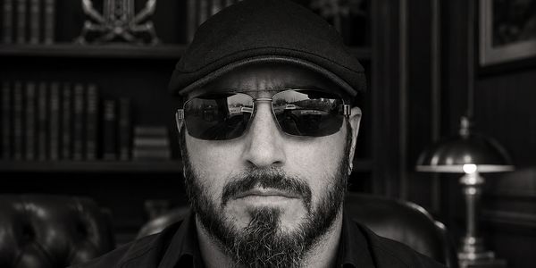 Black and white portrait of a bearded man wearing sunglasses and a flat cap.