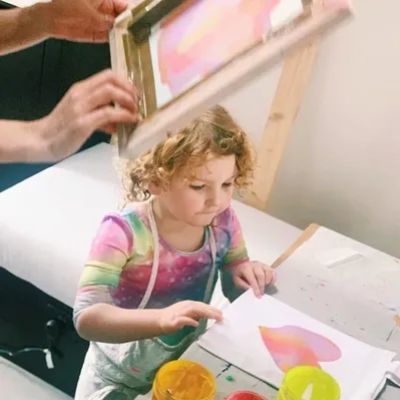 Child in colourful clothes and art apron learns how to screen print love heart bag at art party