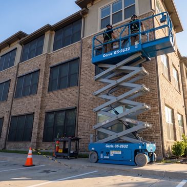 Two workers on a blue scissor lift next to a brick building.
