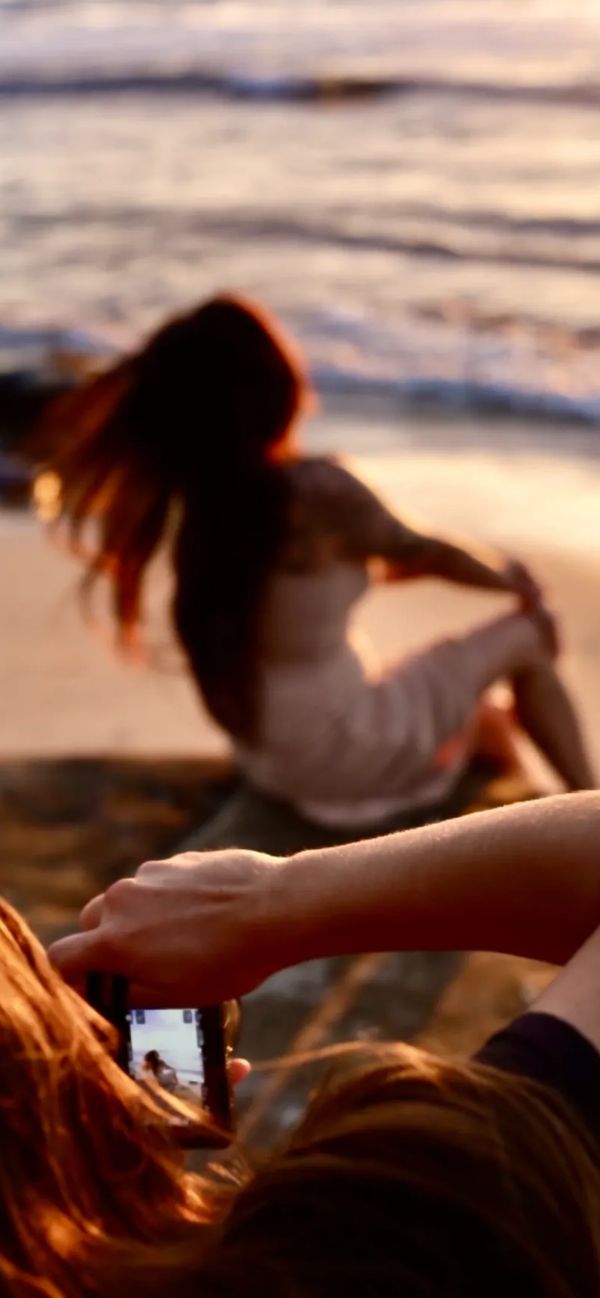 Person taking a photo of a woman sitting by the beach during sunset.