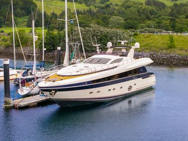 A luxurious yacht docked beside several sailboats in a scenic marina with green hills.