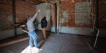Two men inspect exposed pipes and brick walls in a construction site room.