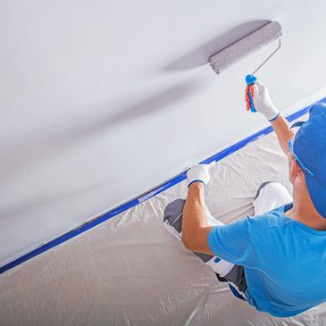 Man painting a wall with a roller while wearing a blue cap and gloves.