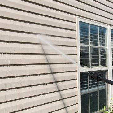 Pressure washing a beige house exterior near windows.