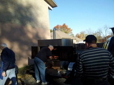 Men grilling meat outdoors using a large smoker on a sunny day.