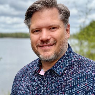 Smiling man with salt-and-pepper hair outdoors near a lake.