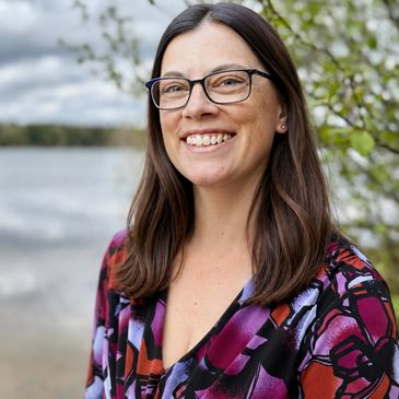 Smiling woman with glasses outdoors by a lake, wearing a colorful patterned top.