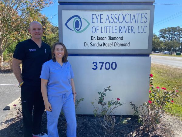Two eye doctors standing beside their clinic sign outdoors on a sunny day.