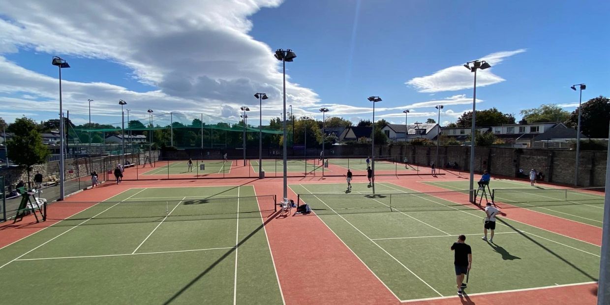 Several people playing tennis on outdoor courts under a blue sky.