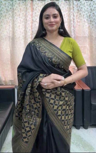 Woman in black and gold saree with green blouse, smiling indoors.