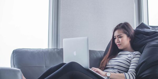 Woman using a laptop while relaxing on a couch near large windows.