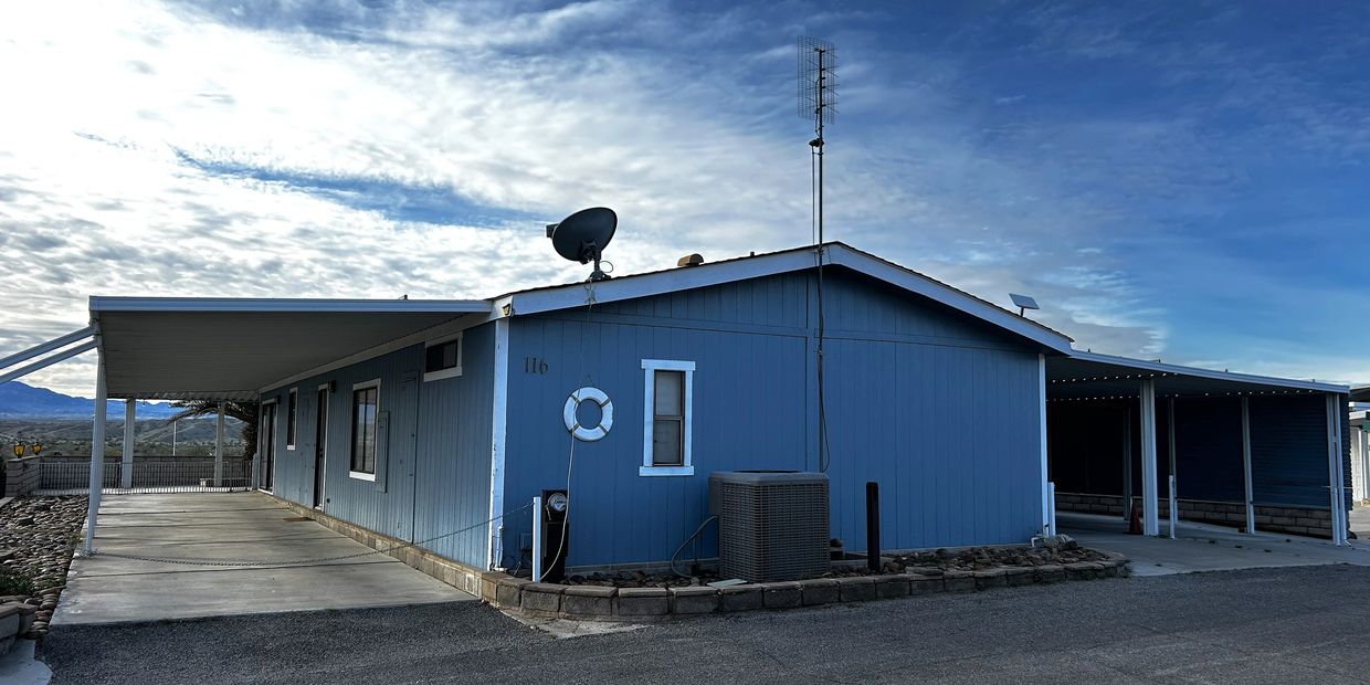 Blue manufactured home with dual covered carports under a cloudy sky.