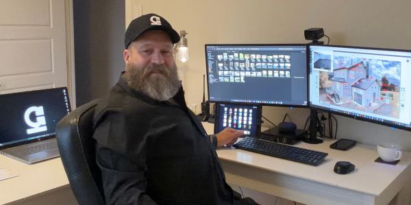 Bearded man with cap working on dual monitors at a desk.
