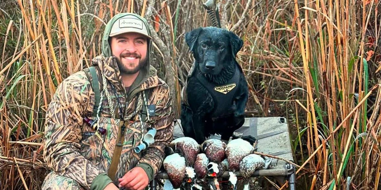 A hunter in camouflage with his black dog and a catch of ducks in a marsh.