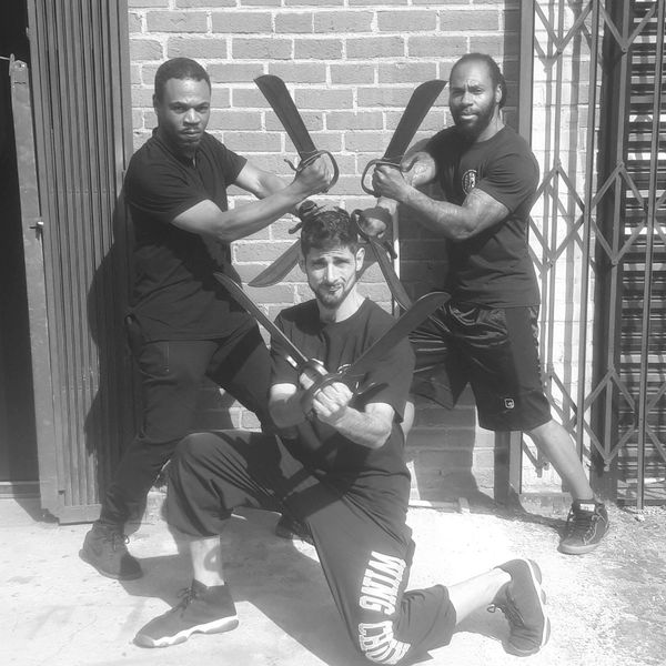 Three men posing with large curved swords outside against a brick wall.