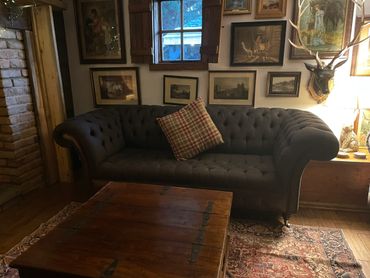 A cozy living room with a dark tufted sofa and plaid pillow under a window and vintage paintings.