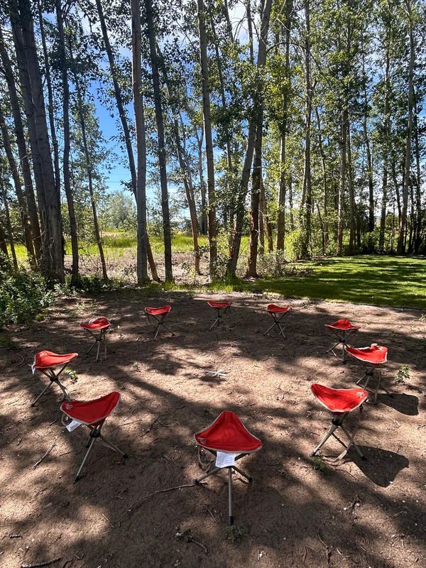 Red folding stools arranged in a circle outdoors under tall trees.