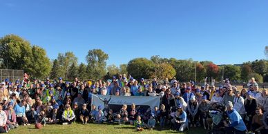 Large group gathered outdoors on a sunny day holding a banner.