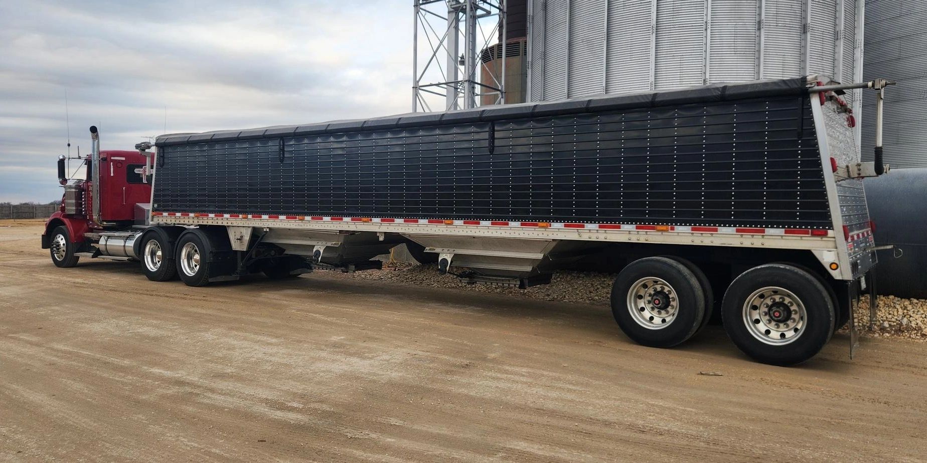 A red semi-truck with a black covered trailer parked on a dirt road.