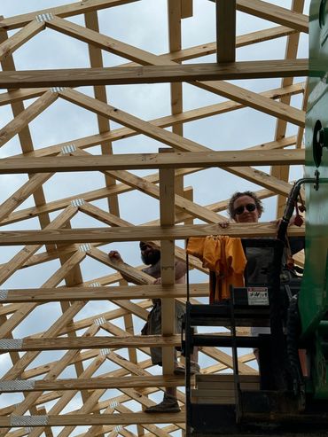 Two construction workers standing among wooden roof trusses under a cloudy sky.