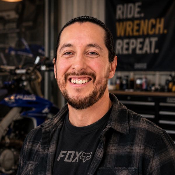 Smiling man with a beard in a workshop with a motorcycle and tools.