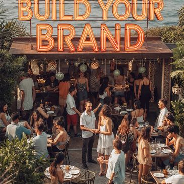People socializing outdoors at a venue under a neon sign saying 'BUILD YOUR BRAND'.
