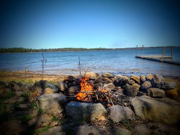 A campfire roars beside a calm lake and wooden dock under a clear blue sky.
