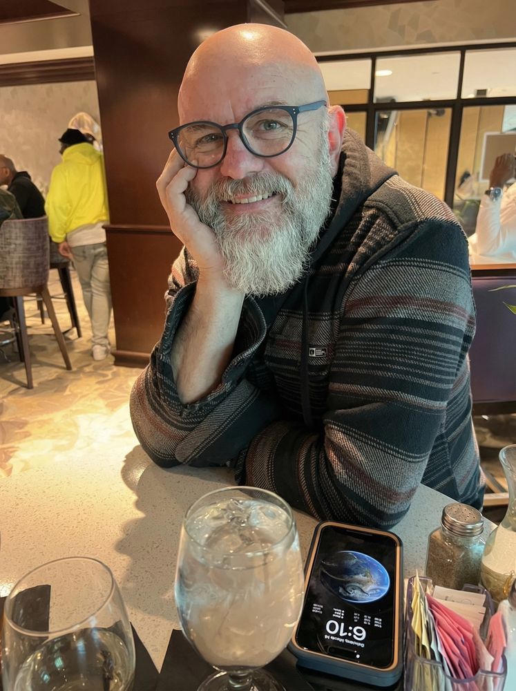 Smiling bearded man in glasses sitting at a table with drinks and phone.